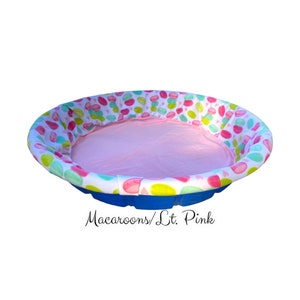 May include: A blue plastic kiddie pool with a pink and white patterned fabric liner. The liner has a pink, green, and white macaroon design. The text "Macaroons/Lt. Pink" is visible below the pool.