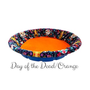 May include: A blue and black pet bed with a Day of the Dead print and an orange cushion. The bed is round and has a scalloped edge.