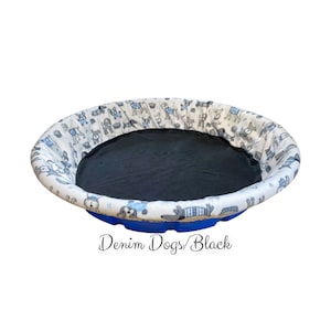 May include: A blue and white dog-themed pet bed with a black fleece liner. The bed is round and has a raised edge. "Denim Dogs/Black" is written below the bed.