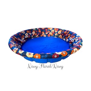 May include: A round pet bed with a navy blue interior and a floral patterned exterior. The floral pattern features orange, yellow, and white flowers on a navy blue background. The bed's base is a solid navy blue. Text on the image reads "Navy Floral/Navy".