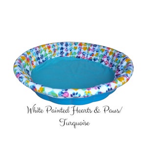 May include: A turquoise dog bed with a white rim and a colorful print of hearts and paw prints. The text "White Painted Hearts & Paws/ Turquoise" is printed below the bed.