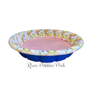 May include: A blue plastic kiddie pool with a pink and white patterned fabric liner. The liner has a pink fleece lining. The text "River Pebbles/Pink" is printed below the pool.