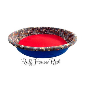 May include: A blue and gray dog bed with a red cushion. The bed is round and has a pattern of dogs on the fabric. The text "Ruff House/Red" is printed below the bed.
