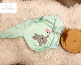 Könnte beinhalten: Ein hellgrüner Pullover mit einem grauen Hasen und einem rosa Schmetterling, die auf der Vorderseite gestickt sind. Der Pullover hat einen Rundhalsausschnitt und lange Ärmel.