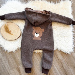 Könnte beinhalten: Brauner Baby-Overall mit Kapuze und einem Bären-Gesicht-Applikation auf der Vorderseite. Der Overall hat eine Tasche und braune Bündchen an Ärmeln und Knöcheln. Das Outfit ist aus weichem, strukturiertem Material.