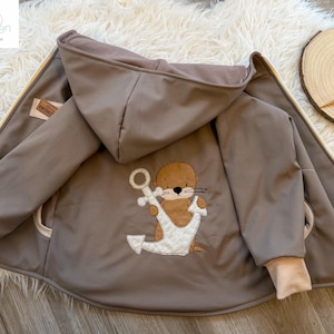 Könnte beinhalten: Braune Kapuzenjacke mit Reißverschluss und beigefarbenen Bündchen. Auf der Rückseite der Jacke ist ein Otter mit Anker gestickt. Die Jacke ist offen und zeigt ein beigefarbenes Innenfutter. Die Jacke liegt auf einer weißen, flauschigen Oberfläche.