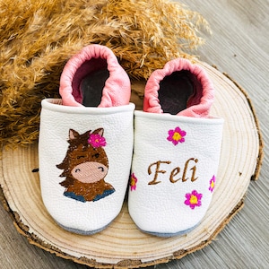 Op de afbeelding: Witte leren babyschoentjes met roze bies. E&eacute;n schoen heeft een geborduurd paardenmotief en de andere heeft de naam "FELI" geborduurd.