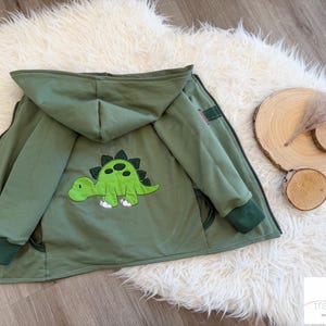 Könnte beinhalten: Olivgrüne Kapuzenjacke mit einem Dinosaurier-Applikation auf dem Rücken. Die Jacke hat einen Reißverschluss, dunkelgrüne Bündchen und eine passende Kapuze. Der Dinosaurier ist grün mit schwarzen Punkten und weißen Füßen. Die Jacke wird offen auf einer weißen, flauschigen Oberfläche präsentiert.