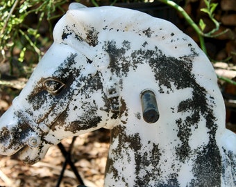 Puede incluir: Un caballo de balancín blanco con salpicaduras de pintura negra, que muestra signos de desgaste. El caballo tiene una brida de cuero negro y un mango de madera.