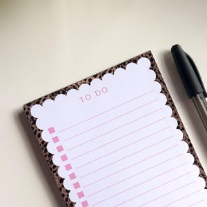 Puede incluir: Un bloc de notas rectangular con un centro blanco festoneado, líneas horizontales rosas y casillas de verificación rosas. El borde tiene un estampado de leopardo marrón y negro. Un bolígrafo negro está colocado junto al bloc de notas.
