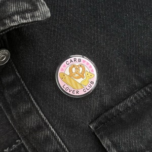 Puede incluir: Un botón rosa y blanco con el texto "Carb Lover Club" y una imagen de dibujos animados de un pretzel y una baguette.