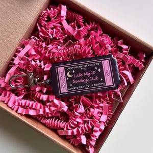 May include: A novelty keychain in a cardboard box filled with pink paper shreds. The keychain is a ticket design with the text "Lifetime Membership to the Late Night Reading Club" and "Always Past Your Bedtime".