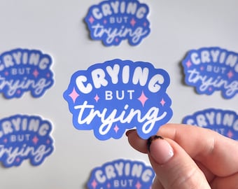 May include: A set of six blue and pink stickers with the text "Crying but trying" in a white, bubbly font. The stickers have pink glitter accents.