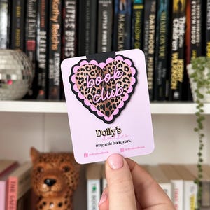 Puede incluir: Un marcapáginas magnético rosa con un diseño de corazón de estampado de leopardo y las palabras "book club". El marcapáginas tiene un borde festoneado y el texto "Dolly's" y "magnetic bookmark" en la parte inferior.