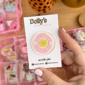 May include: A pink and yellow acrylic pin with the text "Everything will be ok" and a smiley face. The pin is on a white card with the text "Dolly's doodles" and "acrylic pin".