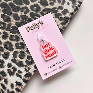 Peut inclure: Un charm transparent pour Kindle avec l'inscription "hot girls read" en lettres rouges. Le charm est fixé à une carte rose avec le texte "kindle charm" et des identifiants de réseaux sociaux. Le fond présente un motif léopard.