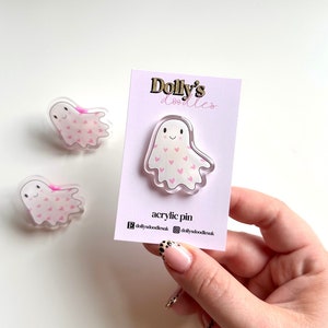 May include: Clear acrylic ghost pin with pink heart details, displayed on a pink card. The card reads "Dolly's doodles" and "acrylic pin". Two more ghost pins are also visible, all with a smiling face.