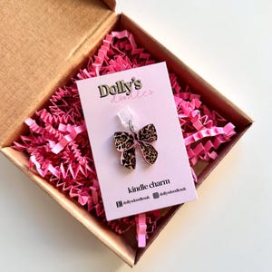 May include: A leopard print bow-shaped charm for a Kindle, presented in a cardboard box with pink paper filler. The charm is attached to a pink card with the text "Dolly's Doodles" and "kindle charm".