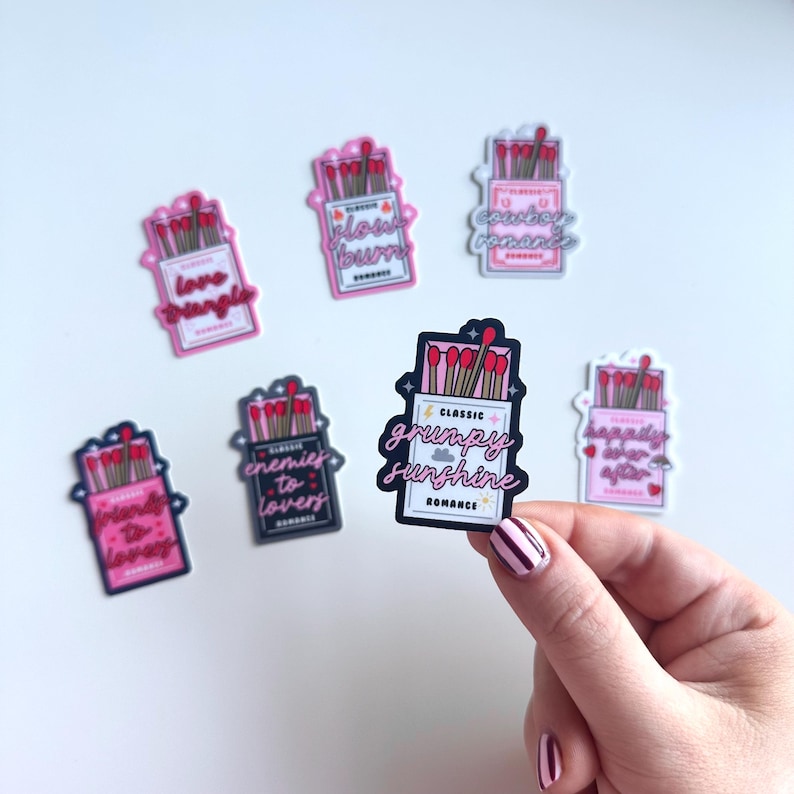 May include: Six pink and black matchbox stickers with different romantic tropes. The stickers feature red matchsticks and white text. The tropes include "Love Triangle", "Slow Burn", "Grumpy Sunshine", "Enemies to Lovers", "Happily Ever After", and "Courtship Romance".