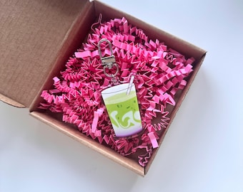 May include: A clear acrylic keychain featuring a matcha latte design with a white cup, green matcha, and a straw. The keychain is attached to a silver clasp and sits inside a cardboard box filled with pink paper shreds.