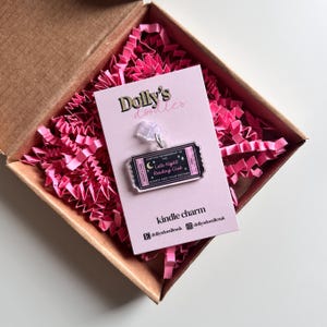 May include: A clear acrylic Kindle charm in a gift box. The charm is shaped like a ticket with the text "Lifetime Membership to the Late Night Reading Club, Always Past Your Bedtime." The box contains pink paper filler.