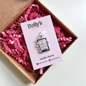 Peut inclure: Un charm en acrylique transparent en forme de liseuse Kindle, avec le texte "This is my emotional support Kindle". Le charm est présenté sur une carte rose avec le texte "Dolly's doodles" dans une boîte marron avec du papier rose.