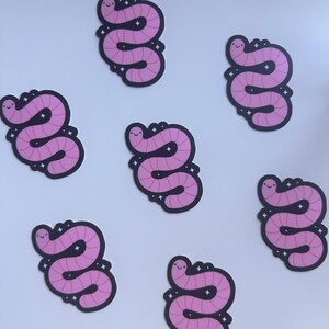 May include: Several stickers featuring a cartoon worm design. The worms are pink with a smiling face and a dark outline. Each sticker has a black border with small white star accents. The stickers are scattered on a white background.