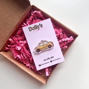 Puede incluir: Un pin acrílico amarillo con forma de taxi de Nueva York, exhibido en una tarjeta rosa dentro de una caja marrón con papel rosa. La tarjeta dice "Dolly's doodles" y "acrylic pin".