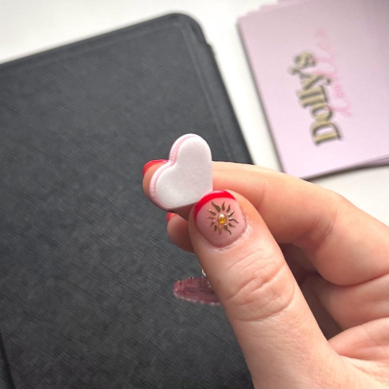 Puede incluir: Un peque&ntilde;o objeto en forma de coraz&oacute;n con un centro blanco y bordes rosas se sostiene en una mano. Las u&ntilde;as est&aacute;n pintadas con una base roja, un dise&ntilde;o de sol dorado y una capa superior transparente. Un fondo negro y una tarjeta rosa con el texto "Dolly's".