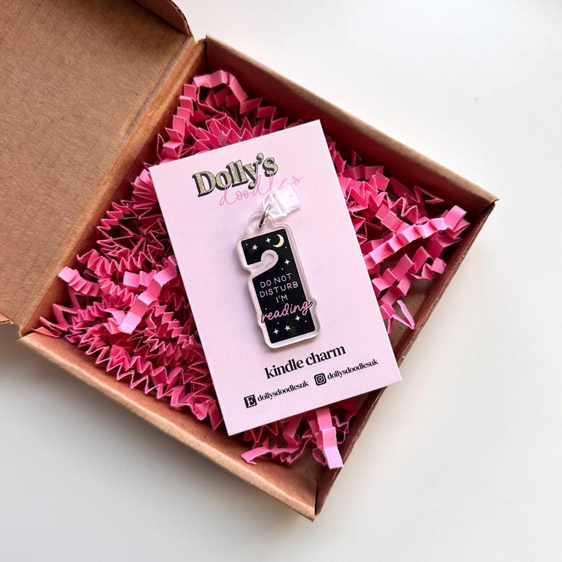 Puede incluir: Un charm de Kindle de acr&iacute;lico transparente con forma de colgador de puerta, con el texto "DO NOT DISTURB I'M reading" y un dise&ntilde;o de cielo nocturno. El charm est&aacute; en una tarjeta rosa dentro de una caja marr&oacute;n con relleno de papel rosa.