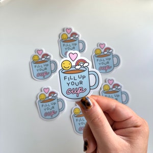 Peut inclure: Un ensemble de six autocollants en forme de tasse à café bleu et blanc avec un arc-en-ciel, un cœur et un smiley. Les autocollants disent "Fill Up Your Cup".
