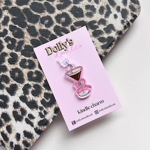 May include: A pink and brown martini charm with a bow detail, attached to a clear earring post, is displayed on a pink card. The card has the text "Dolly's doodles" and "kindle charm". The background is a leopard print pattern.