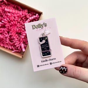 Puede incluir: Un charm Kindle de acr&iacute;lico transparente con forma de colgador de puerta, con un dise&ntilde;o de noche estrellada con una luna creciente y el texto "DO NOT DISTURB I'M reading". El charm est&aacute; unido a una tarjeta rosa con el texto "Dolly's doodles".