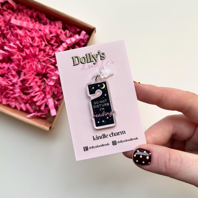 Puede incluir: Un charm Kindle de acr&iacute;lico transparente con forma de colgador de puerta, con un dise&ntilde;o de noche estrellada con una luna creciente y el texto "DO NOT DISTURB I'M reading". El charm est&aacute; unido a una tarjeta rosa con el texto "Dolly's doodles".
