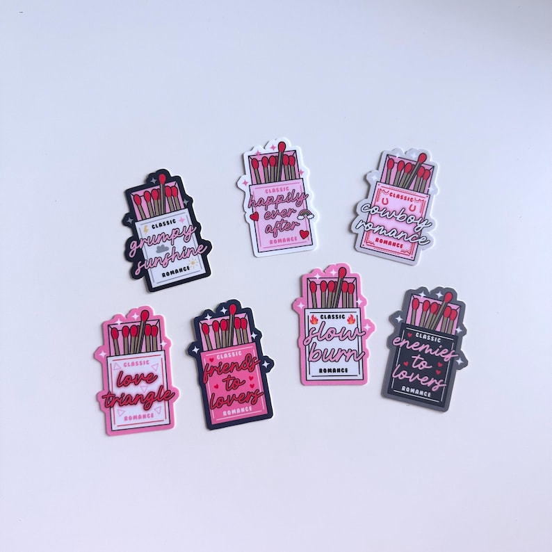 May include: Eight matchbox-style stickers with different romantic themes. Each sticker features a box of matches with red matchsticks and a pink or black background. The text on the stickers includes phrases like "grumpy sunshine", "happily ever after", "cowboy romance", "love triangle", "friends to lovers", "slow burn", and "enemies to lovers".