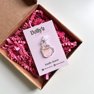 Puede incluir: Un charm de acrílico transparente con forma de taza de té con las palabras "Positive Tea" y un diseño de cara sonriente, corazón y arcoíris. El charm está en una tarjeta rosa dentro de una caja marrón con papel rosa. La tarjeta dice "Dolly's doodles".