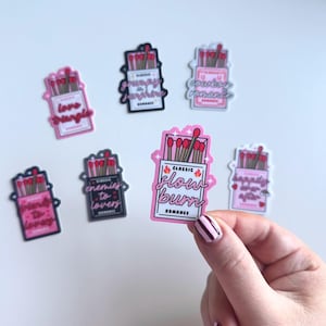 May include: A set of nine pink and black matchbox stickers with different romantic phrases. The stickers feature illustrations of matchsticks and the text "Classic Romance" on each sticker. The phrases include "Love Triangle", "Grumpy & Sunshine", "Cowboy Romance", "Enemies to Lovers", "Happily Ever After", and "Slow Burn".