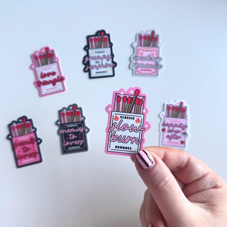 May include: A set of nine pink and black matchbox stickers with different romantic phrases. The stickers feature illustrations of matchsticks and the text "Classic Romance" on each sticker. The phrases include "Love Triangle", "Grumpy & Sunshine", "Cowboy Romance", "Enemies to Lovers", "Happily Ever After", and "Slow Burn".
