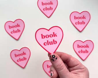 Può includere: Sei adesivi a forma di cuore rosa con il testo "book club" in lettere rosse. Un adesivo è tenuto da una mano.