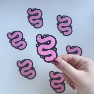 May include: Pink worm stickers with a black outline and a smiling face. The stickers have a glittery design around the worm. Several stickers are scattered on a white surface.