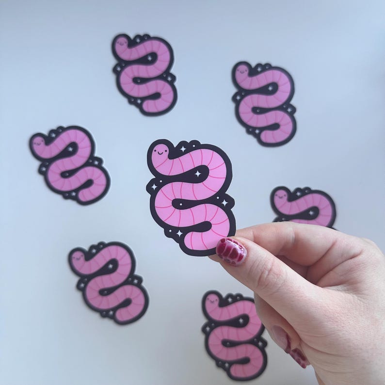May include: Pink worm stickers with a black outline and a smiling face. The stickers have a glittery design around the worm. Several stickers are scattered on a white surface.