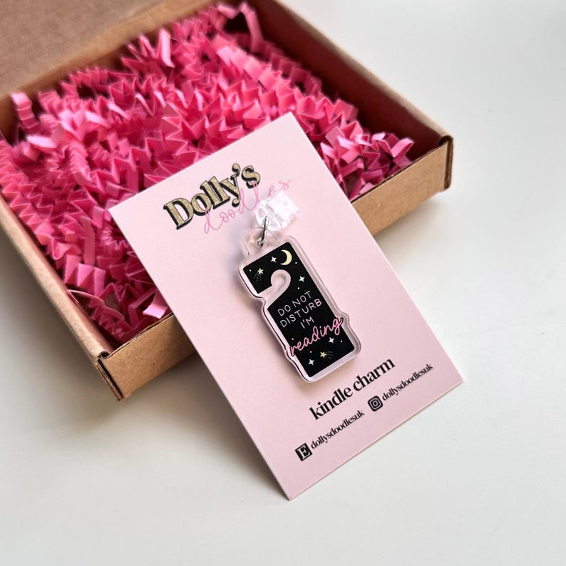 Puede incluir: Un charm de Kindle de acr&iacute;lico transparente con un fondo negro que presenta una luna creciente, estrellas y el texto "DO NOT DISTURB I'M reading". El charm est&aacute; unido a una tarjeta rosa con el logo de Dolly's doodles.
