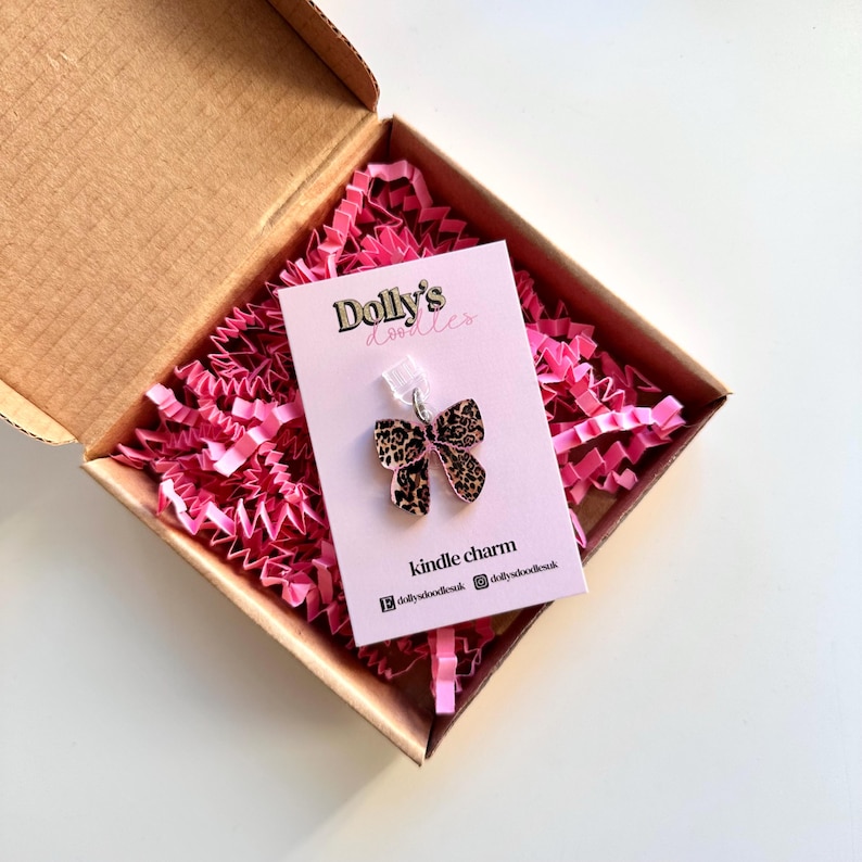 May include: A leopard print bow-shaped charm for a Kindle, presented in a cardboard box with pink paper filler. The charm is attached to a pink card with the text "Dolly's Doodles" and "kindle charm".