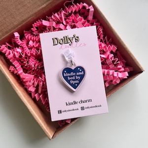 May include: A clear acrylic heart-shaped charm with the text "kindle and bed by 9pm" in a navy blue heart design. The charm is attached to a pink card with the text "Dolly's doodles" and "kindle charm". The charm is packaged in a brown box with pink paper.