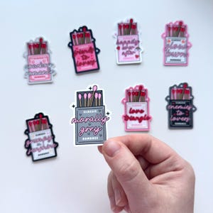 May include: Set of ten pink and gray matchbox stickers with various romantic phrases, including "Cowboy Romance", "Friends to Lovers", "Happily Ever After", "Classic Morally Grey Romance", "Love Triangle", "Enemies to Lovers", "Grumpy Sunshine", "Flowers Burn".