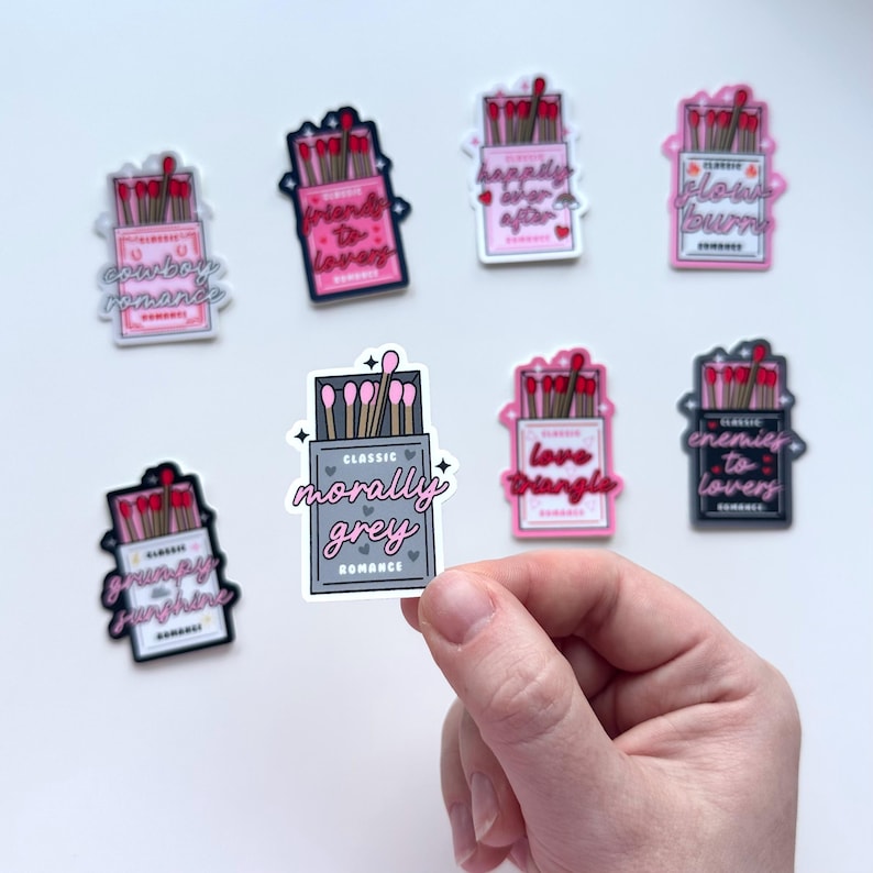 May include: Set of ten pink and gray matchbox stickers with various romantic phrases, including "Cowboy Romance", "Friends to Lovers", "Happily Ever After", "Classic Morally Grey Romance", "Love Triangle", "Enemies to Lovers", "Grumpy Sunshine", "Flowers Burn".