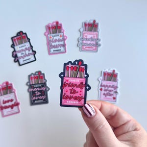 May include: A set of ten pink and white matchbox stickers with different romantic phrases. The stickers feature a box of matches with red matchsticks and a pink label with a romantic phrase. The phrases include "Friends to Lovers", "Grumpy Sunshine", "Enemies to Lovers", "Glow Burn", "Country Romance", "Happily Ever After", "Love Triangle", and "Classic Romance".