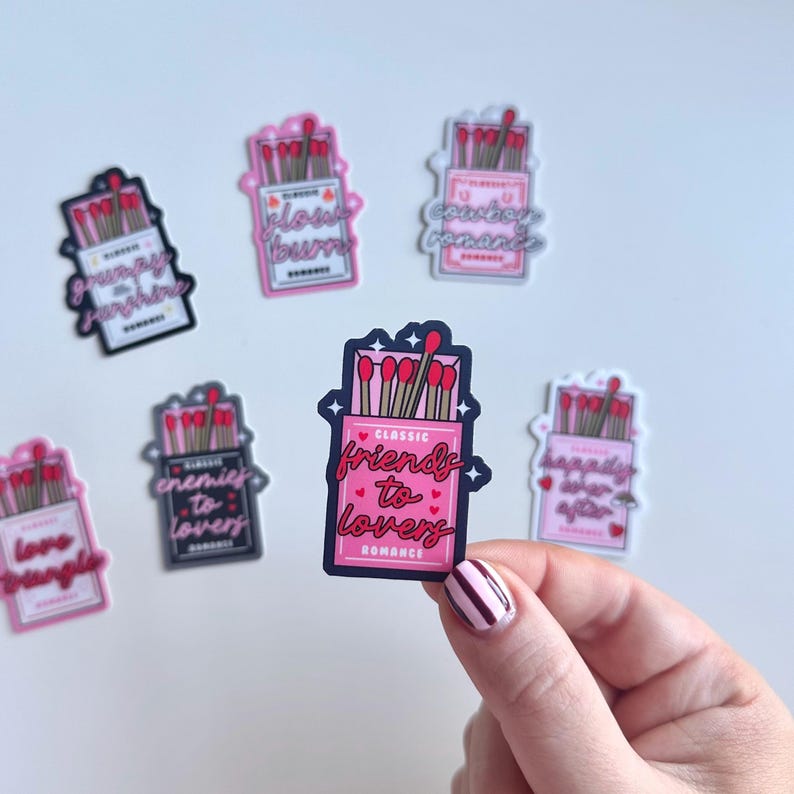 May include: A set of ten pink and white matchbox stickers with different romantic phrases. The stickers feature a box of matches with red matchsticks and a pink label with a romantic phrase. The phrases include "Friends to Lovers", "Grumpy Sunshine", "Enemies to Lovers", "Glow Burn", "Country Romance", "Happily Ever After", "Love Triangle", and "Classic Romance".