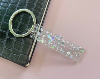 May include: A clear keychain with silver glitter and a white heart charm. The keychain has the text "Minding My Own Small Business" printed on it.