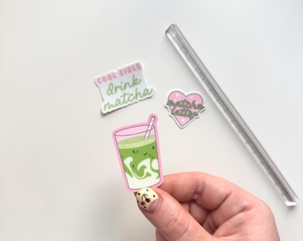 May include: A hand holding a sticker of a matcha latte in a pink-rimmed glass with a straw. Other stickers include a heart-shaped one that says "matcha latte" and one that says "cool girls drink matcha". A clear, rectangular straw is also present.